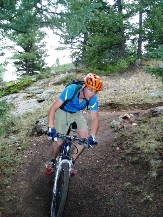 A cyclist wearing an orange helmet and a blue shirt rides a mountain bike along a dirt trail surrounded by trees and rocky terrain. The cyclist is focused and leaning forward, navigating a bend in the path. Happy Jack Recreation Area mountain bike trail.