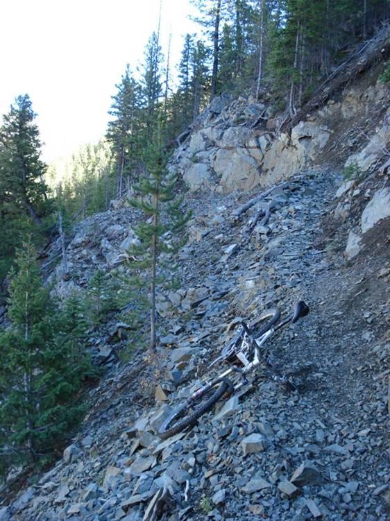 A mountain bike resting on a rocky, uneven trail surrounded by pine trees in a forested area. Sunlight filters through the trees in the background, illuminating the steep incline and scattered rocks along the path. Rock Creek mountain bike trail.
