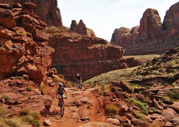 Two mountain bikers riding on a dirt trail through a rugged red rock landscape, surrounded by towering cliffs and natural formations under a clear sky. Amasa Back Trail mountain bike trail.