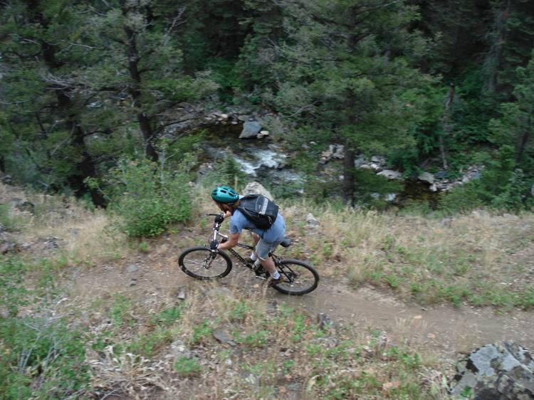 A mountain biker navigating a dirt trail surrounded by trees and greenery, with a small stream visible in the background. The rider is wearing a blue helmet and a backpack, pedaling uphill on a rocky path. Rock Creek mountain bike trail.