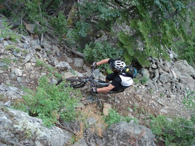 Aerial view of a mountain biker navigating a rocky trail surrounded by greenery and trees. The biker, wearing a helmet and a black and white outfit, is focused on maneuvering over uneven terrain. Rock Creek mountain bike trail.