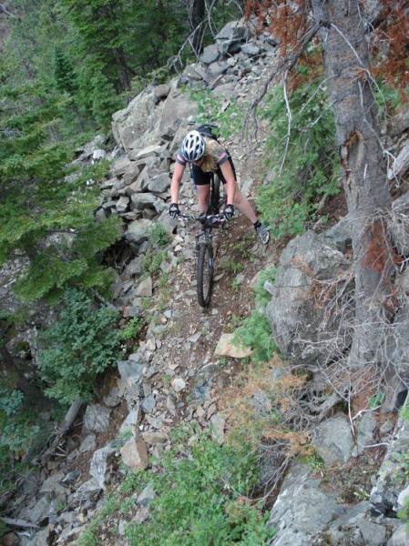 A mountain biker navigating a rocky trail surrounded by green trees and rugged terrain. The rider is leaning forward on the bike, focused on balancing on the narrow path. Rock Creek mountain bike trail.