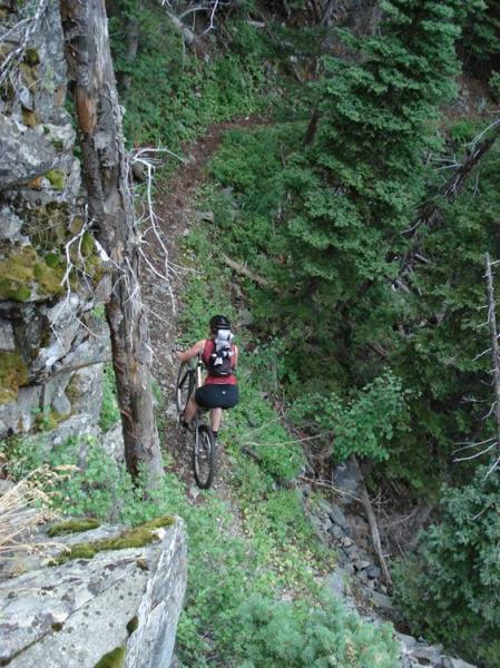 A mountain biker navigating a narrow dirt trail surrounded by dense greenery and rocky terrain. The biker is seen from above, riding along the edge of a steep slope with trees and foliage in the background. Rock Creek mountain bike trail.