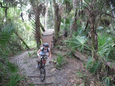 A mountain biker navigating a rugged trail surrounded by lush greenery and tropical plants, with a wooden bridge visible in the background. North Port Mountain Bike Trails mountain bike trail.