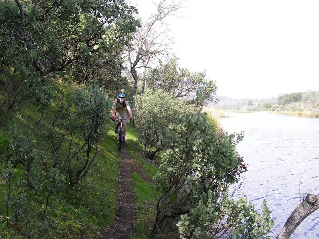 A mountain biker riding along a narrow trail lined with greenery, next to a calm body of water. The scene captures the natural landscape with trees and bushes around the pathway, showcasing an outdoor recreational activity. Pachecho Falls mountain bike trail.