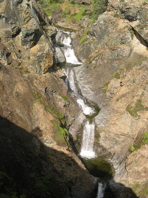 Aerial view of a multi-tiered waterfall cascading down a rocky cliff, surrounded by lush greenery and rugged terrain. The water flows over smooth stones, creating a serene natural scene. Pachecho Falls mountain bike trail.