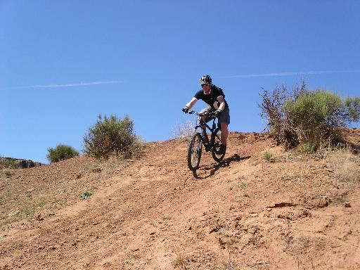 A person riding a mountain bike down a dirt slope, surrounded by sparse vegetation under a clear blue sky. Bearclaw Poppy mountain bike trail.