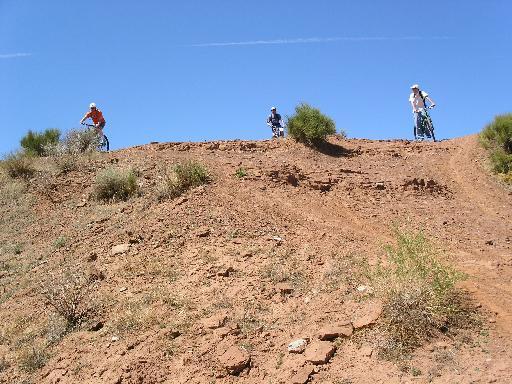 Three mountain bikers riding on a dirt trail at the top of a hill with a clear blue sky. The landscape features sparse vegetation and rocky terrain. Bearclaw Poppy mountain bike trail.