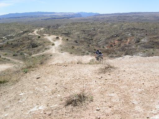 A motorcyclist riding on a dirt path through an arid landscape with rolling hills, sparse vegetation, and a blue sky in the background. Bearclaw Poppy mountain bike trail.