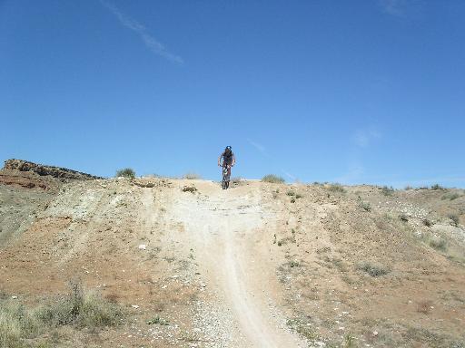A person riding a mountain bike up a dirt trail on a hillside under a clear blue sky. The landscape features arid terrain with sparse vegetation and rocky formations in the background. Bearclaw Poppy mountain bike trail.