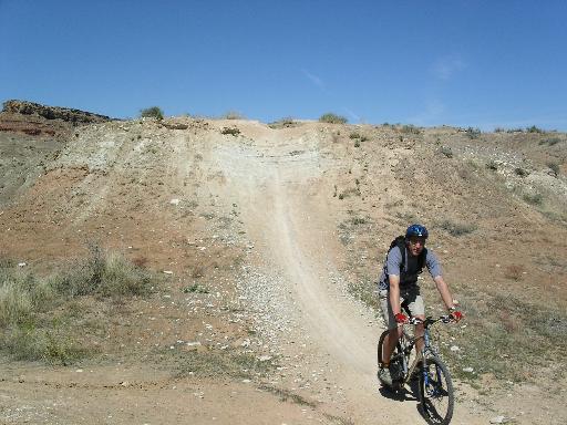 A person riding a mountain bike on a dirt trail near a hill, with a clear blue sky in the background. The terrain is rocky and arid, with sparse vegetation. Bearclaw Poppy mountain bike trail.