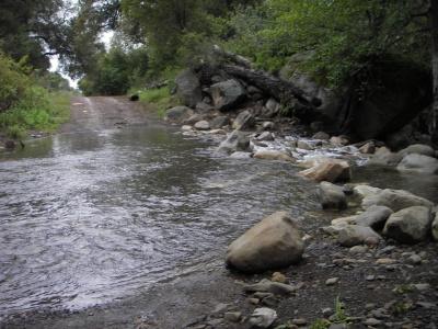 A serene landscape featuring a shallow stream flowing over smooth rocks, with a dirt road visible in the background. Lush greenery surrounds the water, and large boulders can be seen along the stream
