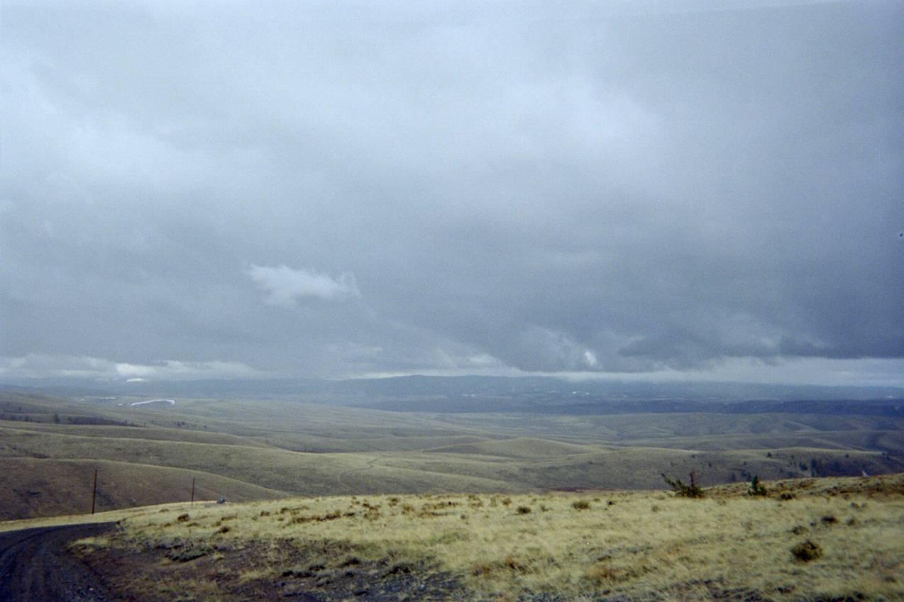 A panoramic view of rolling hills and valleys under an overcast sky. The landscape is mostly dry with sparse grass and a dirt road winding through the foreground, leading to the distant horizon. The scene conveys a sense of solitude and natural beauty amid gray skies. Jelm Mountain Road mountain bike trail.