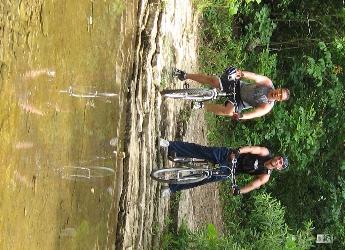 Two cyclists posed on bicycles near a shallow stream, with a rocky shoreline and lush greenery in the background. The water reflects their images, creating a scenic outdoor setting. Busiek State Forest mountain bike trail.