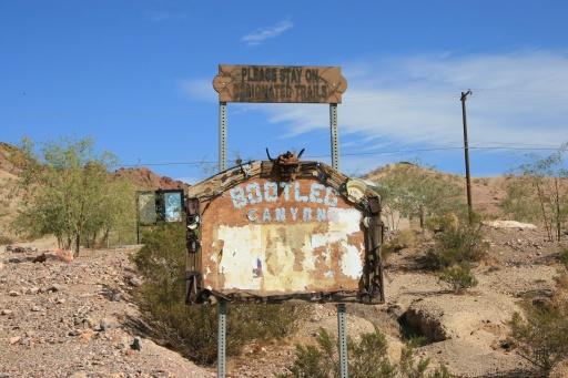 An old, weathered sign that reads "Please Stay On Designated Trails" above a partially obscured sign for "Bootleg Canyon." The sign is rusty and peeling, set against a backdrop of rocky terrain and sparse vegetation under a clear blue sky. Bootleg Canyon mountain bike trail.