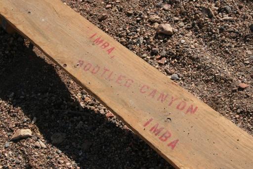Wooden plank with red markings that read "IMBA" and "BOOTLEG CANYON" on a gravel surface. Bootleg Canyon mountain bike trail.