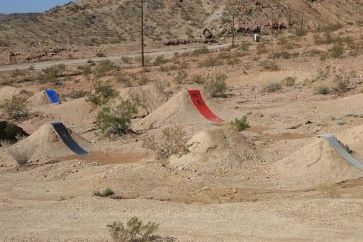 A dirt jump park featuring several mounds with colorful ramps, set in a dry, rocky landscape. The mounds are designed for biking and include ramps painted in blue, red, and gray. Sparse vegetation and distant hills create the backdrop. Bootleg Canyon mountain bike trail.