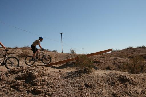 A mountain biker riding over a wooden ramp in a desert landscape, with a clear blue sky and telephone poles in the background. Bootleg Canyon mountain bike trail.