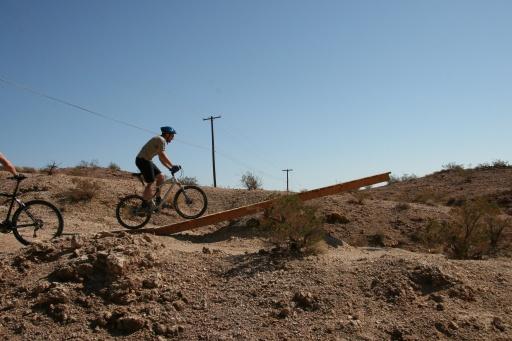 A mountain biker wearing a helmet navigates a wooden ramp on a rocky terrain under a clear blue sky. In the background, there are power lines and sparse vegetation, typical of a desert landscape. Bootleg Canyon mountain bike trail.