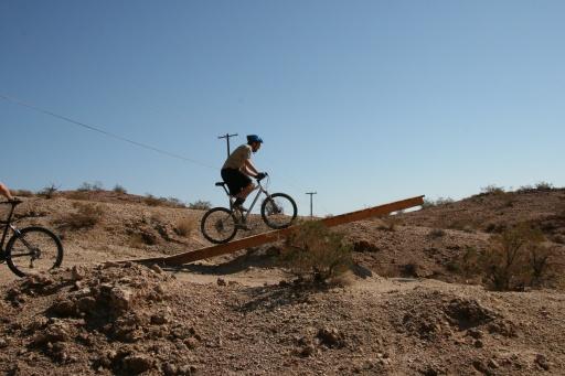 A cyclist riding a mountain bike across a wooden ramp in a rocky desert landscape under a clear blue sky. Bootleg Canyon mountain bike trail.
