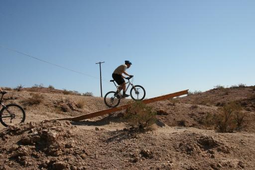 A person riding a mountain bike down a wooden ramp in a desert landscape under a clear blue sky. The scene shows rocky terrain with sparse vegetation and a distant power line. Bootleg Canyon mountain bike trail.