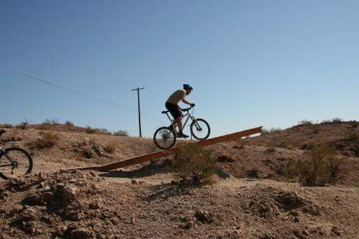 A cyclist riding a mountain bike going up a wooden ramp on a dirt trail, surrounded by rocky terrain and sparse vegetation under a clear blue sky. Bootleg Canyon mountain bike trail.