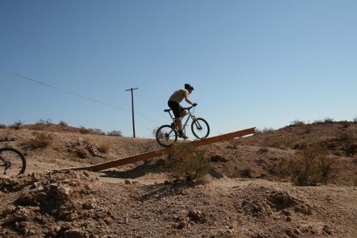 A person riding a mountain bike on a wooden ramp in a rugged, desert landscape under a clear blue sky. Bootleg Canyon mountain bike trail.
