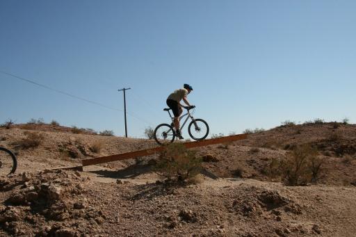 A mountain biker navigating a wooden plank bridge across a rocky terrain under a clear blue sky. Bootleg Canyon mountain bike trail.