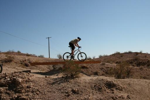 A mountain biker performing a jump on a wooden bridge over rough terrain, under a clear blue sky. The landscape includes hills and sparse vegetation, with a nearby power pole in the background. Bootleg Canyon mountain bike trail.