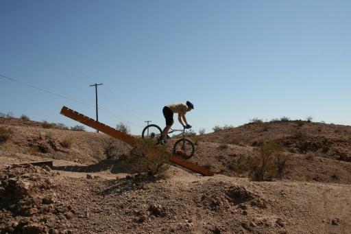 A cyclist performs a trick on a wooden ramp in a desert landscape, with rocky terrain and sparse vegetation under a clear blue sky. Bootleg Canyon mountain bike trail.
