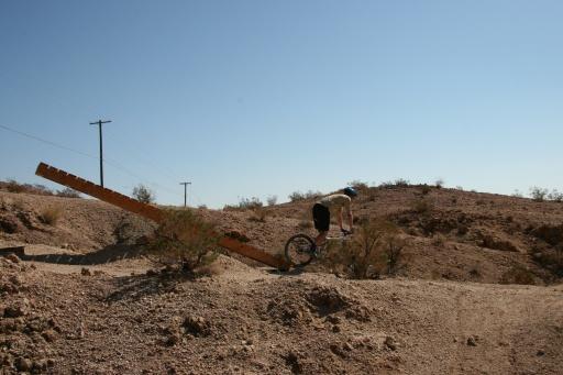 A cyclist navigating a wooden ramp in a desert landscape, with sparse vegetation and power lines in the background under a clear blue sky. Bootleg Canyon mountain bike trail.