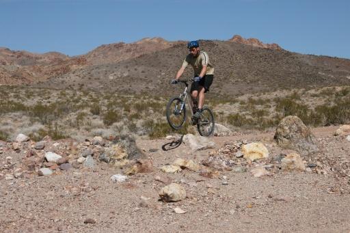 A person riding a mountain bike on rocky terrain in a desert landscape, with mountains in the background and sparse vegetation. Bootleg Canyon mountain bike trail.