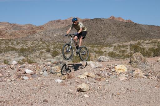 A mountain biker in mid-air performing a jump over a small rock in a desert landscape, surrounded by sparse vegetation and rocky hills in the background under a clear blue sky. Bootleg Canyon mountain bike trail.