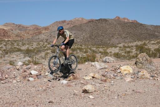 A person riding a mountain bike over rocky terrain in a desert landscape, with mountains in the background. The cyclist is wearing a helmet and is captured mid-ride, navigating through the natural obstacles. Bootleg Canyon mountain bike trail.