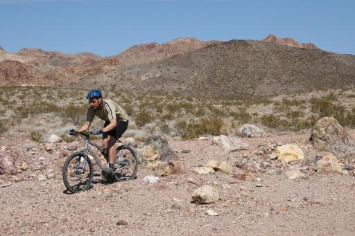 A cyclist wearing a helmet rides a mountain bike across a rocky, arid landscape with colorful hills in the background. The terrain is uneven, featuring scattered rocks and sparse vegetation under a clear blue sky. Bootleg Canyon mountain bike trail.
