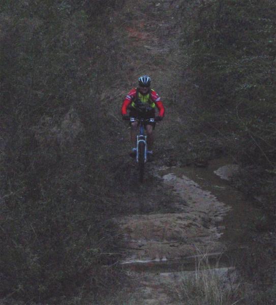 A cyclist riding a mountain bike along a narrow, rugged trail surrounded by dense vegetation and rocky terrain. The scene is dimly lit, suggesting early morning or late evening, with the cyclist wearing a bright red and yellow jersey and a helmet, navigating through a muddy section of the path. UWF Mountain Bike Trails mountain bike trail.