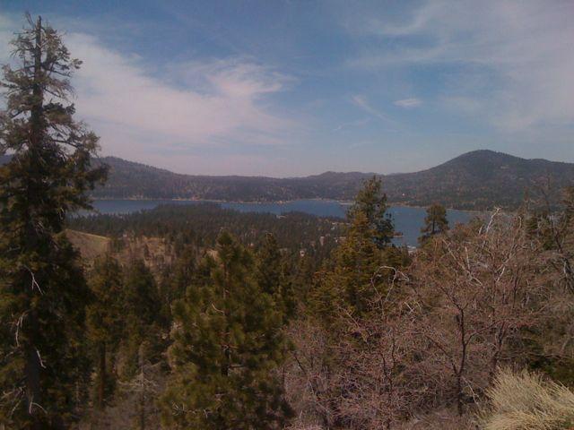A scenic view of a lake surrounded by forested hills under a partly cloudy sky. The foreground features various evergreen trees, while the background showcases the calm blue water of the lake and distant mountains. Pine Knot Trail mountain bike trail.