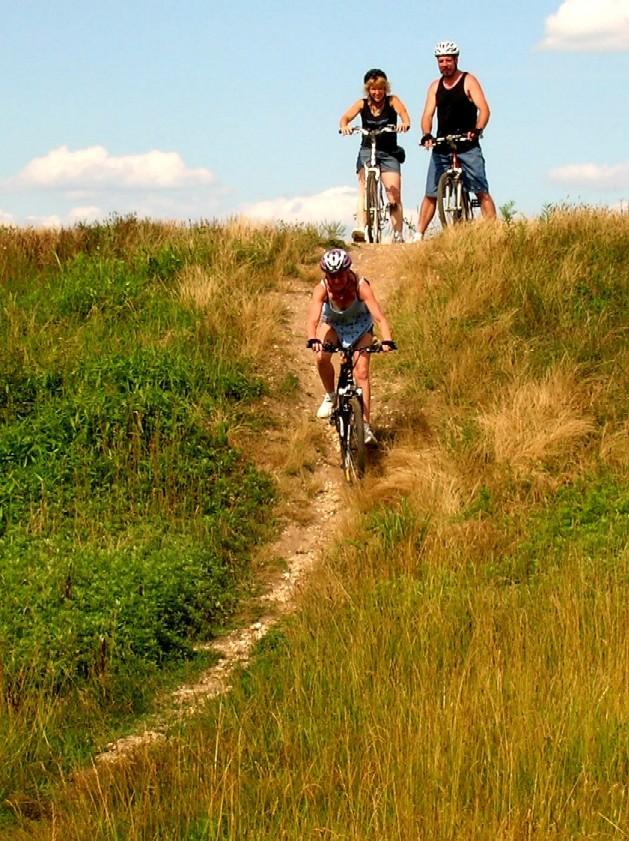 Three people are biking on a grassy hill; one cyclist is riding downhill in the foreground, while two others are visible at the top of the hill, preparing to descend. The sky is clear with a few clouds, and the landscape features lush green grass and a dirt path. Mercer County Park mountain bike trail.