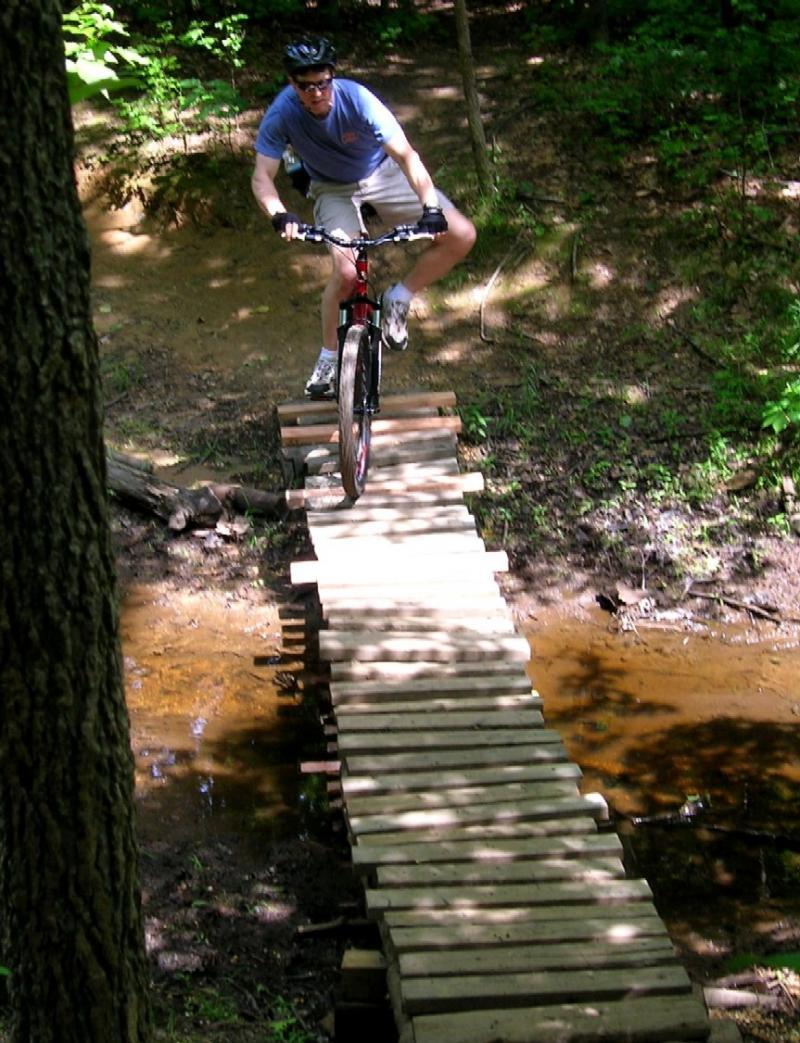A person riding a mountain bike is navigating a wooden plank bridge over a shallow stream in a wooded area. The cyclist is wearing a helmet and sunglasses while focused on maintaining balance as they cross the bridge. Surrounding greenery indicates a sunny day in nature. Mercer County Park mountain bike trail.