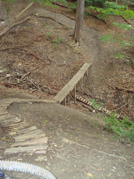 A narrow wooden bridge spans a small ravine in a forested area, surrounded by trees and scattered leaves. The ground slopes down towards the bridge, with a few wooden planks visible on the approach. The scene captures a natural, rustic environment ideal for outdoor activities. Don Valley mountain bike trail.