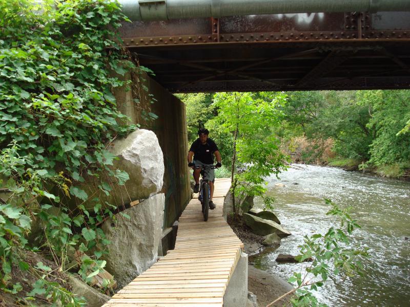 A mountain biker riding along a narrow wooden path that runs beside a river, with lush greenery and large rocks on either side. An overhead metal bridge is visible in the background. Don Valley mountain bike trail.