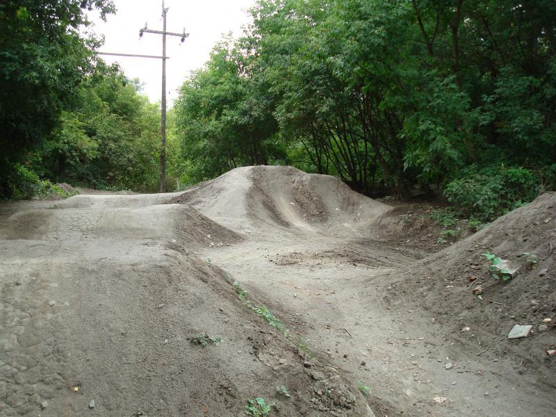 A dirt bike track with various mounds and jumps, surrounded by trees and shrubs, along a path with a utility pole in the background. Don Valley mountain bike trail.