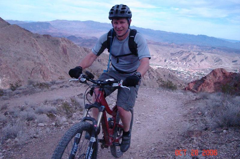 A man in a helmet and athletic attire is riding a mountain bike on a rocky trail in a mountainous landscape. He is smiling and appears to be enjoying the ride, with a view of distant hills and a town below. The scene is set in a natural setting with sparse vegetation and rugged terrain. Bootleg Canyon mountain bike trail.