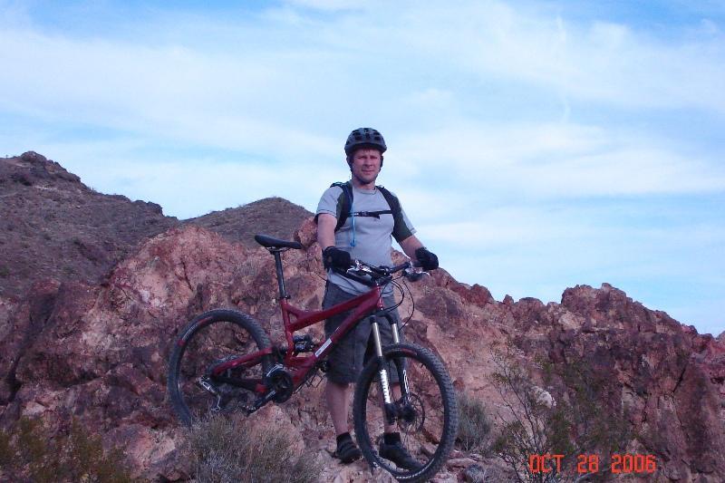 A person standing on rocky terrain with a mountain bike, wearing a helmet and sports attire, against a backdrop of hills and a cloudy sky. Bootleg Canyon mountain bike trail.