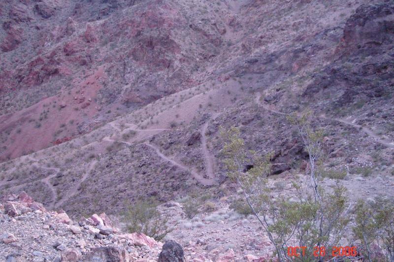 A rugged, mountainous landscape featuring a winding dirt trail descending through a canyon. The terrain is predominantly rocky with patches of sparse vegetation and reddish-brown earth tones, conveying a sense of remote natural beauty. The sky is clear, suggesting a dry environment typical of desert regions. Bootleg Canyon mountain bike trail.