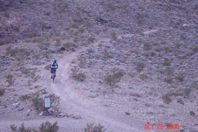 A mountain biker rides along a winding dirt trail through a rocky, desert landscape with sparse vegetation. The scene is captured from a distance, highlighting the rugged terrain and the biker's movement. A sign is visible near the trail. Bootleg Canyon mountain bike trail.