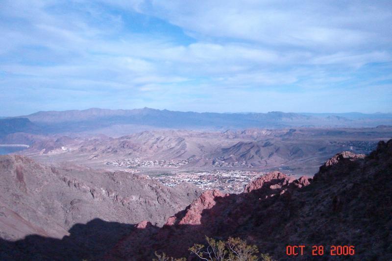 A panoramic view of a mountainous landscape under a partially cloudy sky, showcasing rugged terrain and distant hills, with a small town visible in the valley below. Bootleg Canyon mountain bike trail.