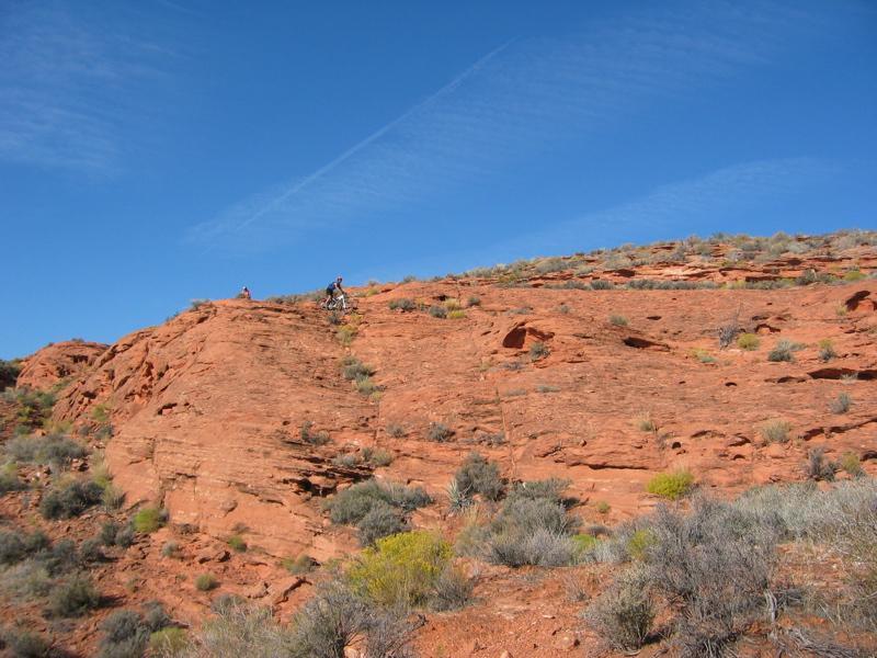 Two people are seen near the top of a red rock formation under a clear blue sky. The rocky terrain is dotted with sparse vegetation, including small shrubs and patches of grass. Church Rocks mountain bike trail.