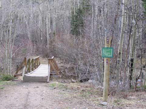 A wooden footbridge spans a small stream, surrounded by dense trees and underbrush, with a green informational sign on a post nearby. The scene is set in a wooded area, indicating a natural hiking or walking trail. Silverdale mountain bike trail.