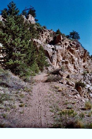 A dirt trail winding through a rocky landscape, surrounded by patches of grass and small trees. In the background, a clear blue sky is visible, and a wooden utility pole can be seen at the top of the cliff. Empire Pass mountain bike trail.